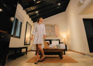 Guest in a white linen dress exploring a minimalist boutique suite at Beldon Hiriketiya, featuring vaulted dark wood ceilings and warm lighting.