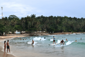 Panoramic view of Hiriketiya Beach with surfers in the lineup and travelers chilling by the turquoise seaside