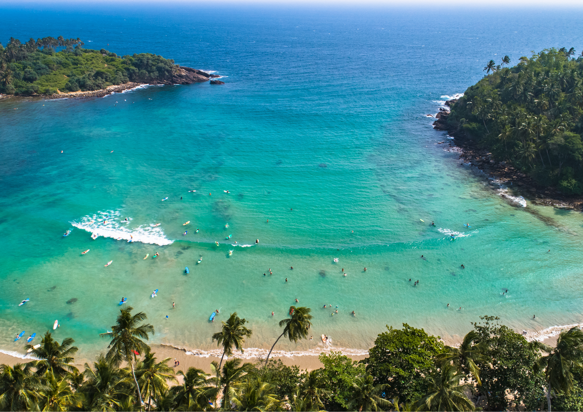 Aerial drone view of Hiriketiya Beach Sri Lanka showing surfers catching waves in the turquoise horseshoe bay surrounded by tropical palm trees.