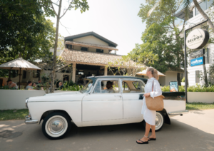 A boutique traveler stands next to a classic white vintage car parked outside Beldon Boutique Hotel in Hiriketiya.