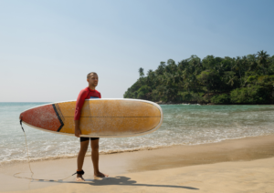 A male surfer holding an orange surfboard on the sandy shoreline of the horseshoe bay while analyzing hiriketiya surfing beginners wave conditions.
